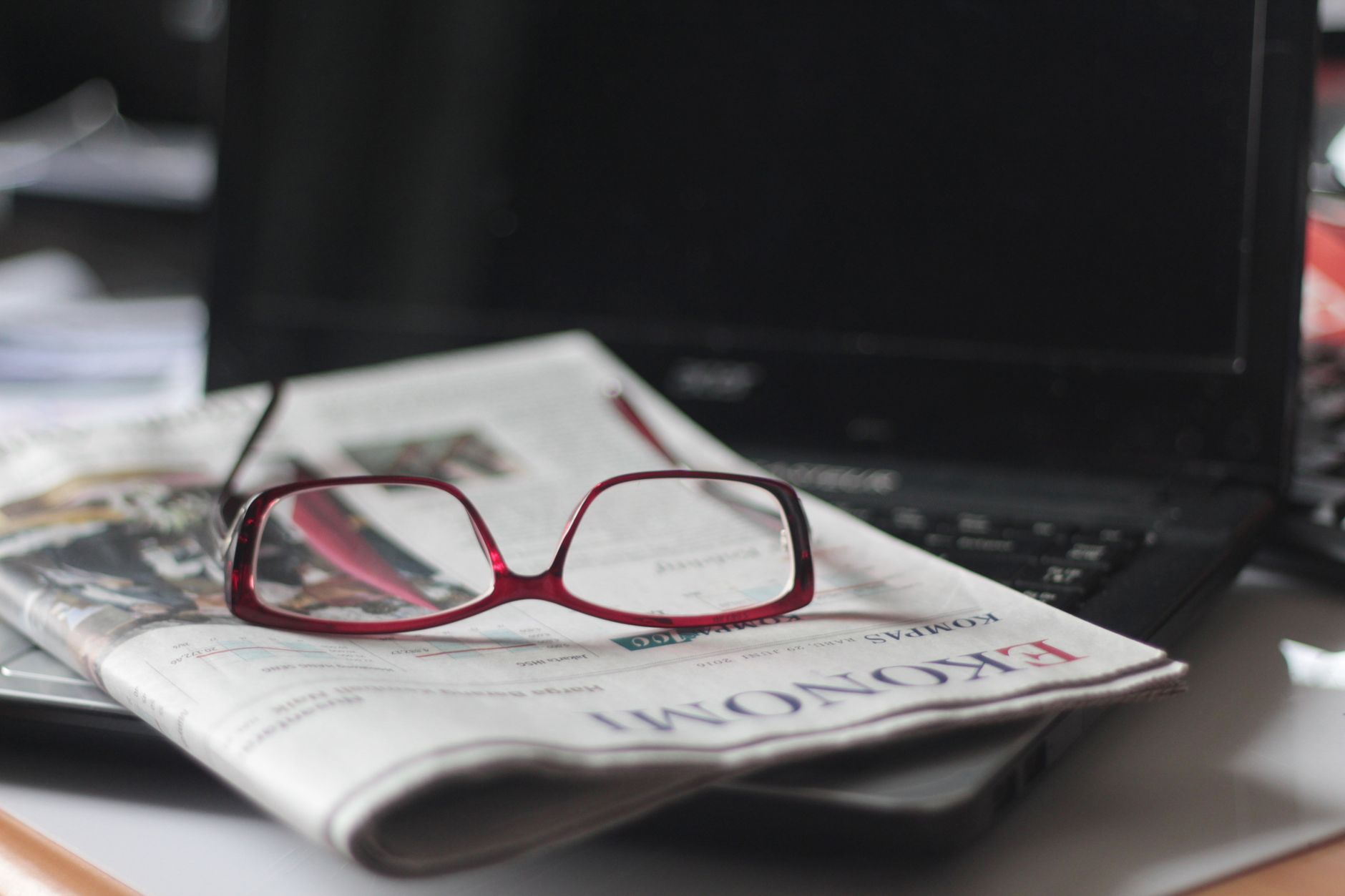 Stack of printed newspapers next to an open laptop displaying a news website in a quiet newsroom